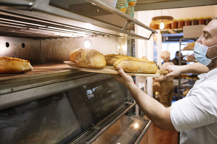 Male chef in protective face mask removing fresh bread from oven in bakery kitchen