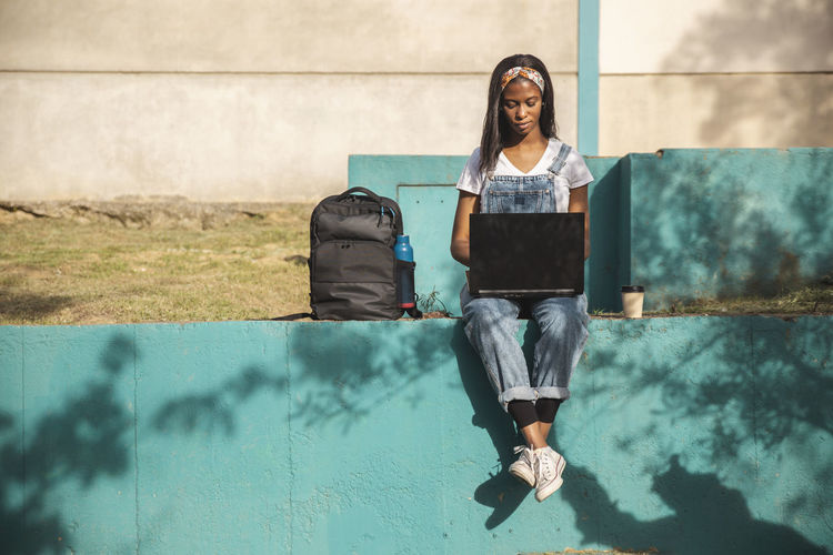 Young freelancer using laptop sitting on wall