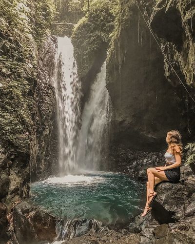 Full length of young woman looking at waterfall while sitting on rock
