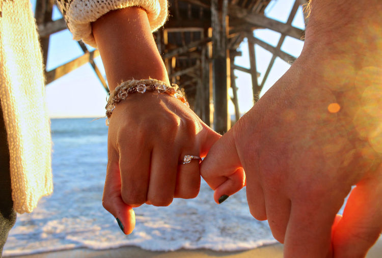 Cropped hand of man and woman holding little finger against sea