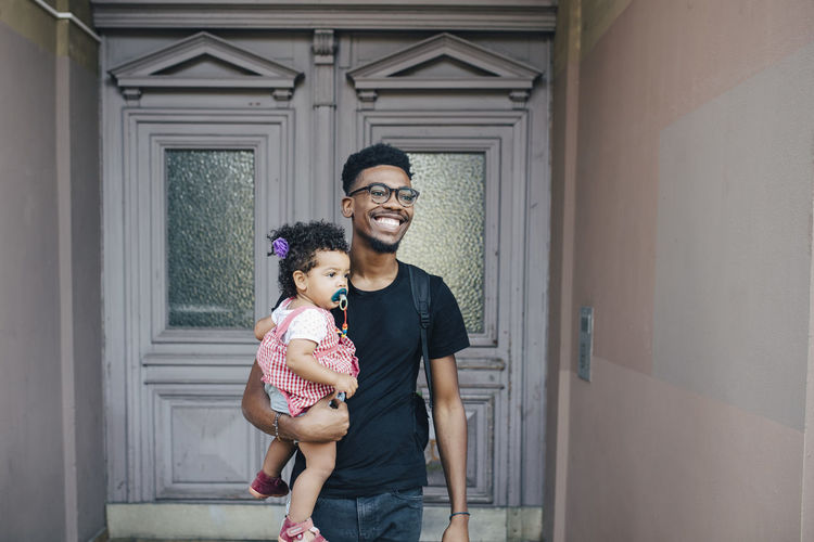 Smiling father looking away while carrying daughter against closed door