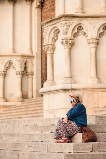 Side view of female tourist in protective mask sitting on stairs near column of medieval stone building and talking on smartphone while resting during sightseeing in cuenca town in spain
