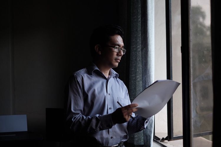 Man reading document while standing by window at home