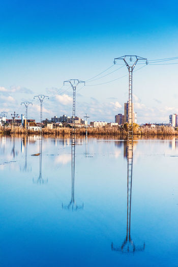 Reflection of electricity pylon in water against blue sky