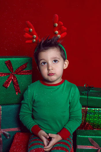 Baby boy in a green t shirt and with deer antlers sits with gifts on a red background