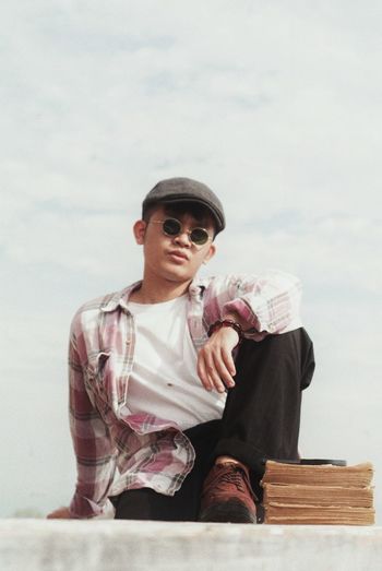 Low angle view of young man with books wearing sunglasses while sitting on retaining wall against sky