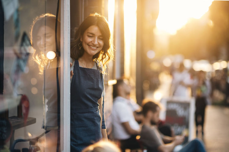Smiling female owner looking down while standing at entrance of deli