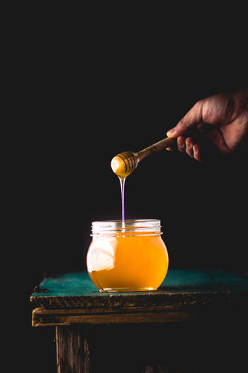 Cropped hand holding honey dipper over glass jar on table against black background