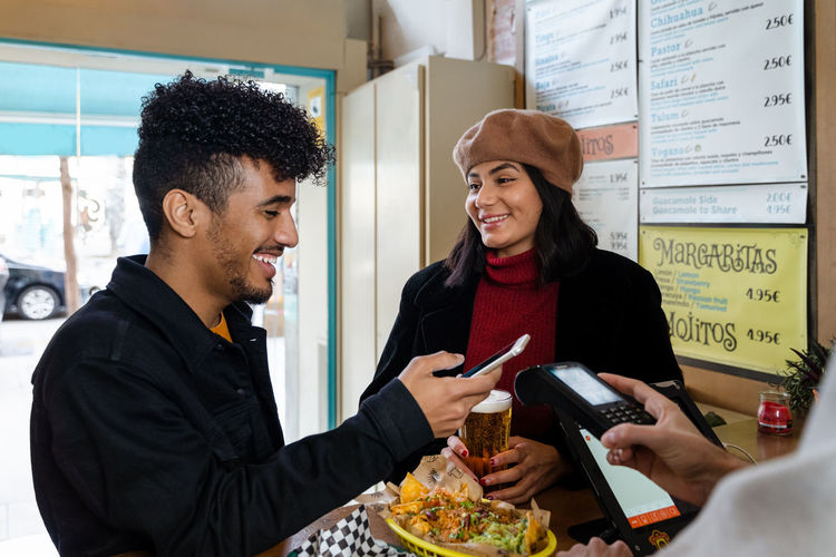 Cheerful young ethnic couple paying for food order with smartphone while standing near counter in light restaurant