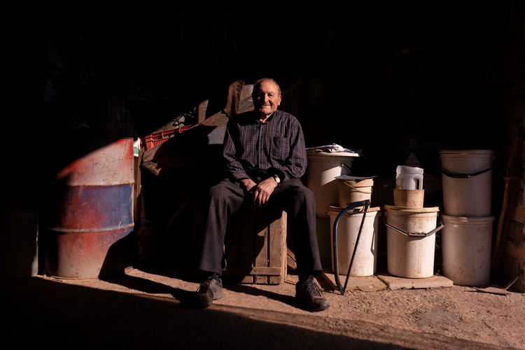 Full body of happy senior male sitting on wooden box in garage among containers in garage at sunlight looking at camera