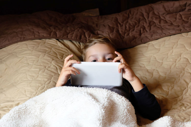 Little boy surfing the net on digital tablet wile relaxing on a bed.