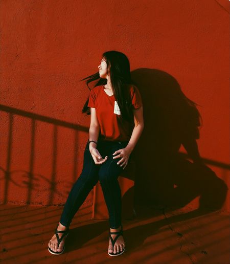 Full length of woman tossing hair while sitting against red wall
