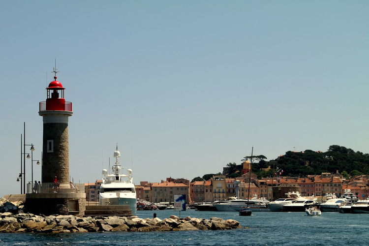 Lighthouse and buildings by boats moored at river against clear sky