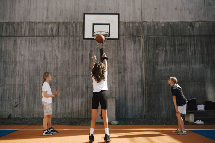 Rear view of girl throwing basketball in hoop while playing with friends at sports court