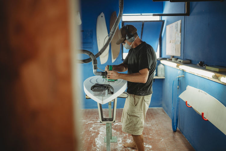 Side view of qualified worker in protective mask handling white surfboard in workshop with blue walls