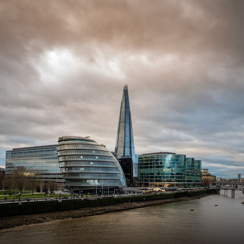 Modern buildings by river against cloudy sky