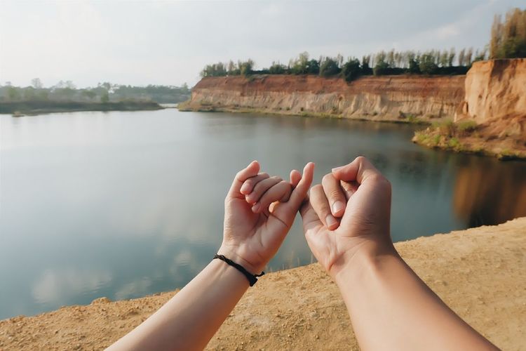 Cropped hands of couple taking pinky promise by lake against sky