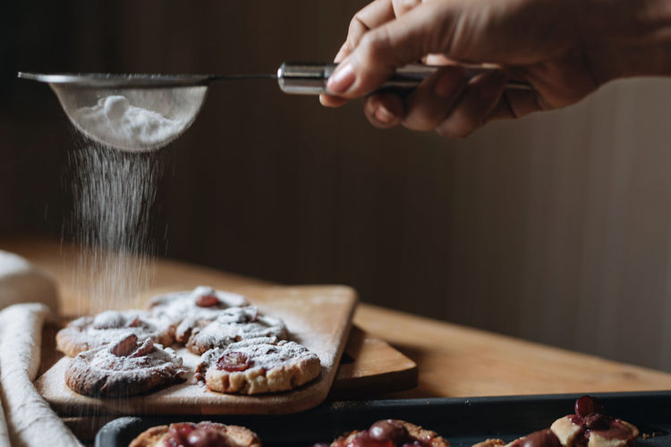 Cropped hand of woman dusting powdered sugar on cookies with strainer on table
