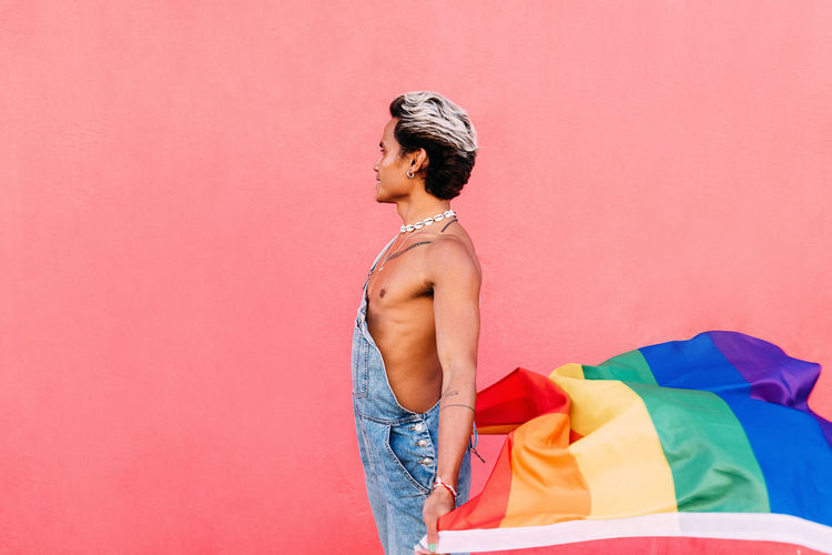 Side view of gay man holding rainbow flag against pink background