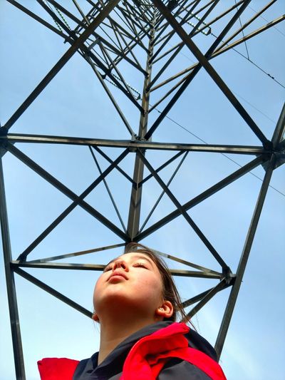 Low angle view of girl standing below electricity pylon against sky