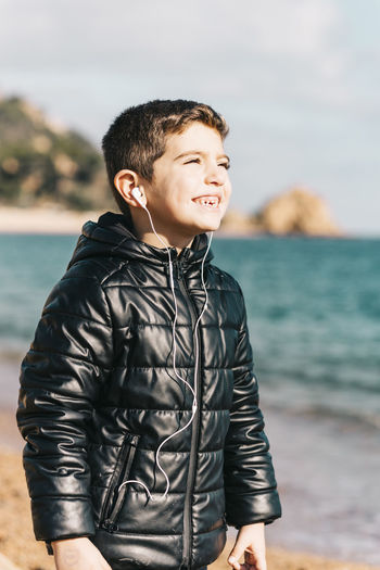 Smiling boy listening music while standing at beach against sky