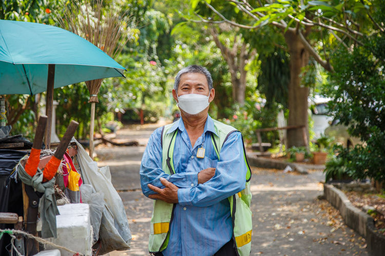 An asian man street cleaner wearing a mask standing with his arm crossed next to an old garbage cart
