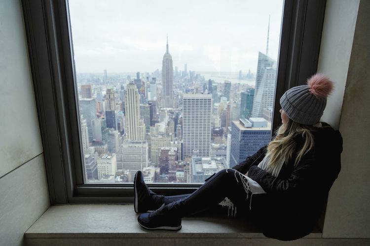 Side view of young woman looking at empire state building through window 