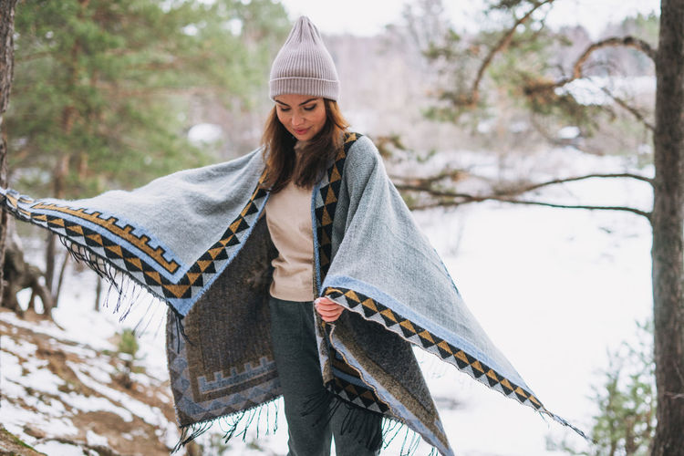 Young brunette beautiful woman in hat and grey poncho in the winter forest