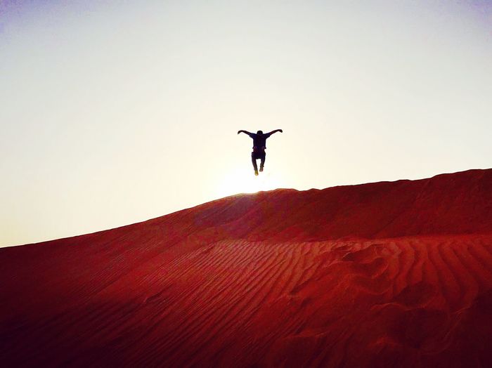 Low angle view of person with arms outstretched jumping over sand in desert against clear sky