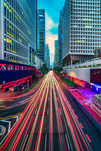 Light trails on city street by buildings against sky