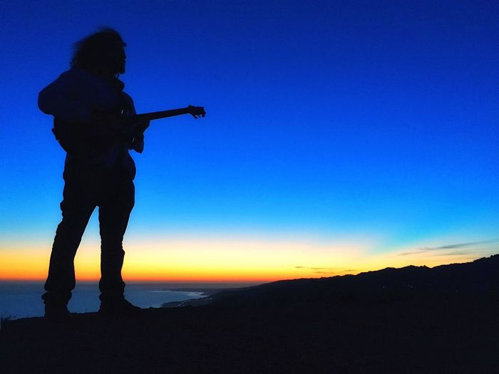 Silhouette guitarist standing at beach against sky during sunset