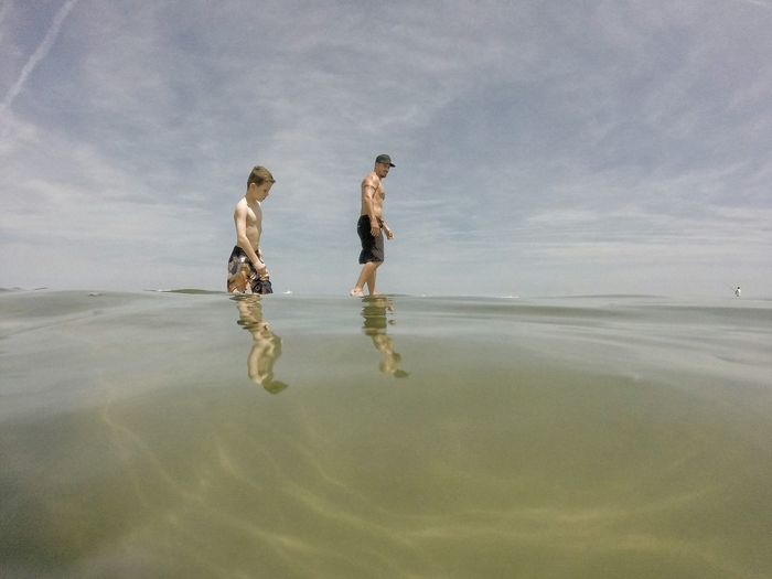 Water level of shirtless father and son in sea against sky