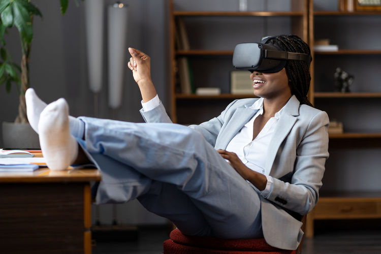 Curious afro businesswoman in vr goggles headset touching 3d virtual objects sit at office workplace