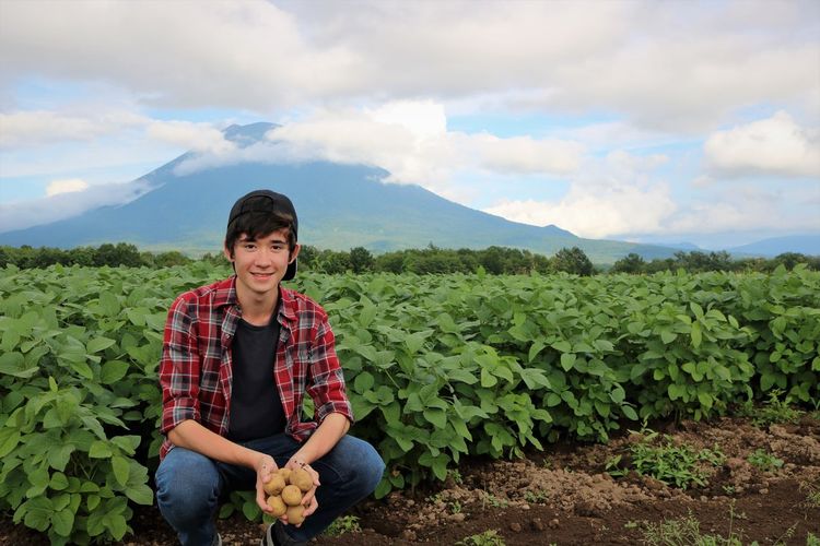 Portrait of smiling teenage boy holding raw potatoes by plants on field against cloudy sky
