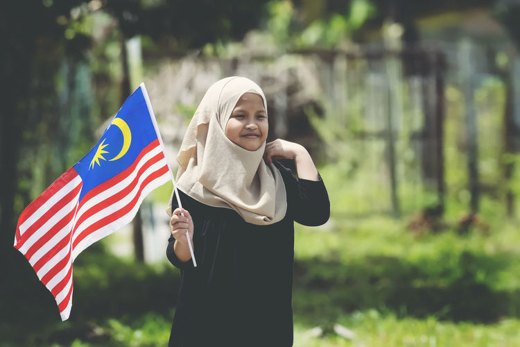 Girl in traditional clothing holding malaysian flag while standing at park
