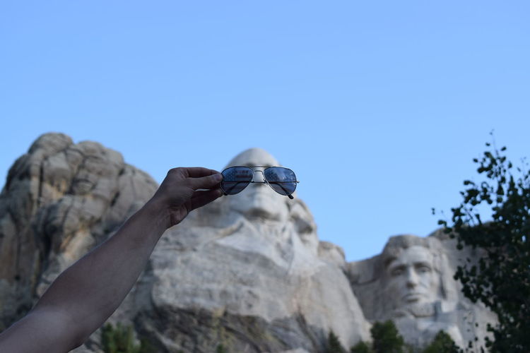 Close-up of hand holding sunglasses on mount rushmore national memorial against sky