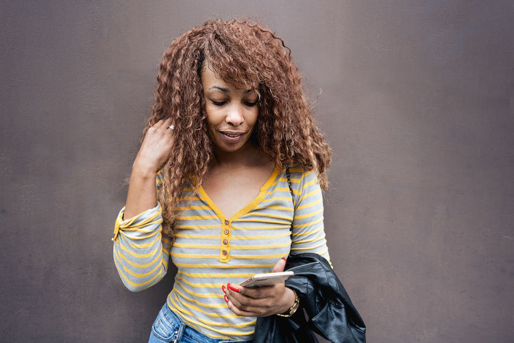 Woman with hand in hair using phone while standing against wall