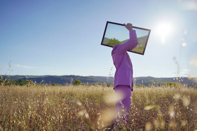 Woman holding mirror standing on field against sky