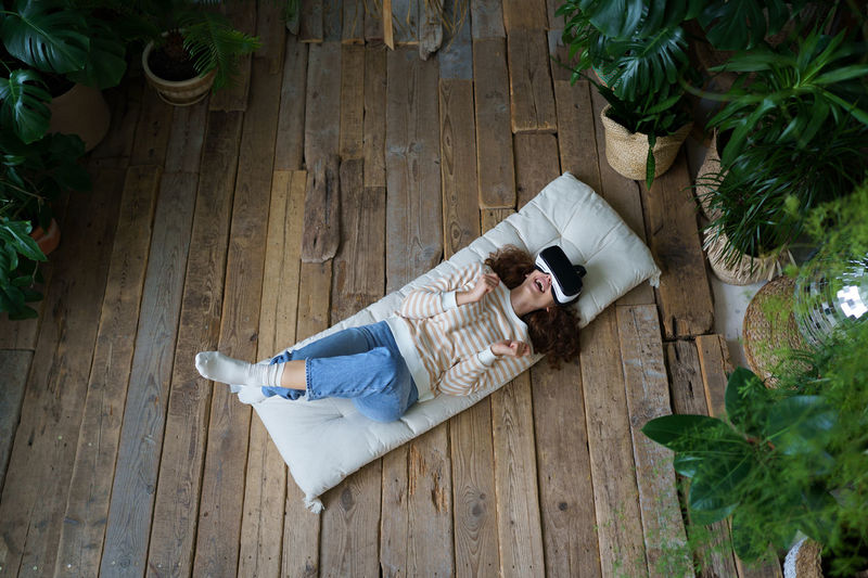 Young excited woman in vr helmet resting in greenhouse, lying on mattress enjoying virtual reality