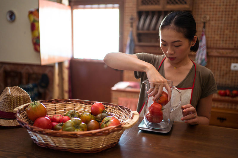 Focused ethnic housewife weighing fresh tomatoes in glass jug on kitchen scale while cooking food at home