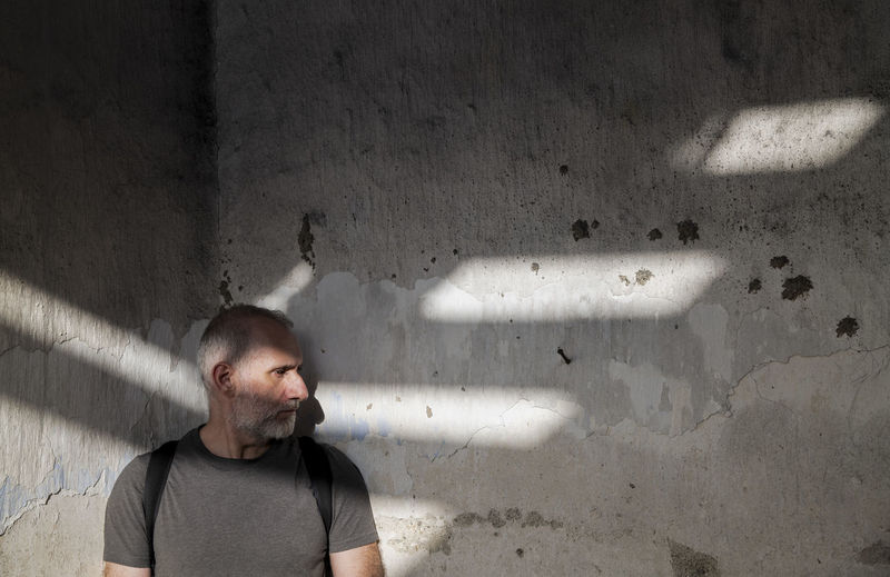 Portrait of adult man looking away while standing on old wall with sunlight and shadow