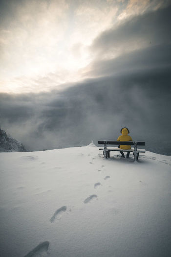 Rear view of mid adult man sitting on bench at snowcapped mountain against sky