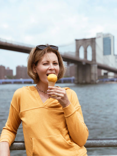 Tourist woman eating  cone of sorbet against background of manhattan city and brooklyn bridge.