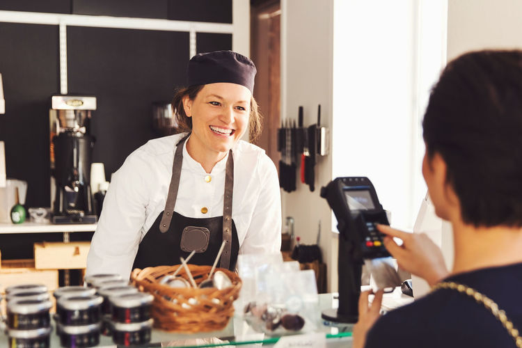 Happy saleswoman looking at female customer entering pin in credit card reader at grocery store