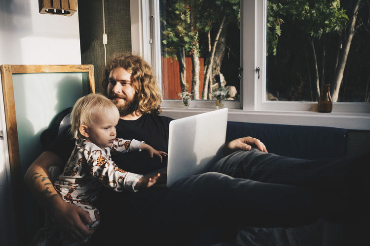 Father with girl holding laptop while sitting by window at home