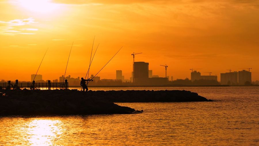 Silhouette factory by sea against orange sky