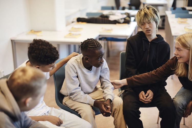 Blond teenage girl consoling boy sitting amidst male friends in classroom during group therapy