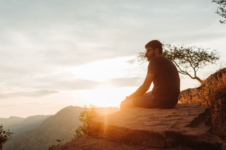 Side view of man sitting on cliff against sky during sunrise