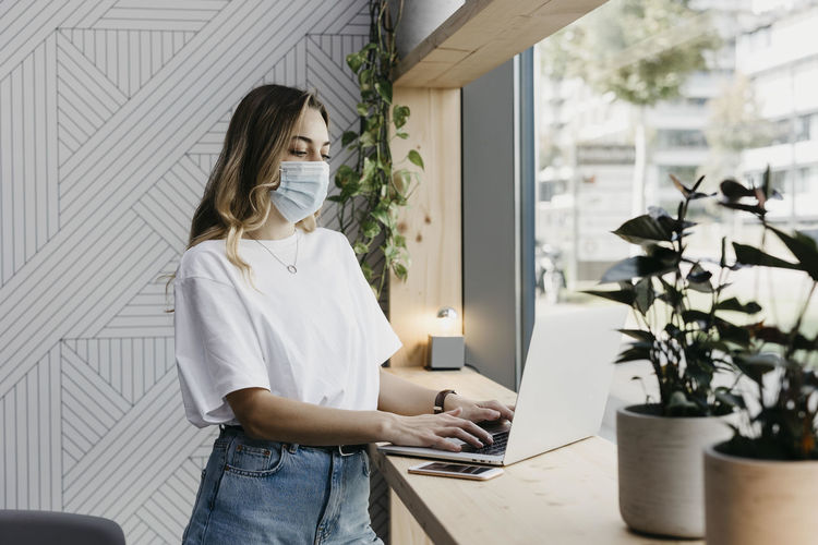 Woman wearing protective face mask using laptop while standing in cafe during covid-19