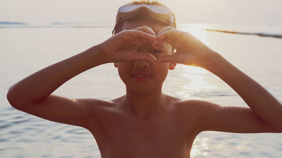Portrait of smiling boy making heart shape against sea during sunset